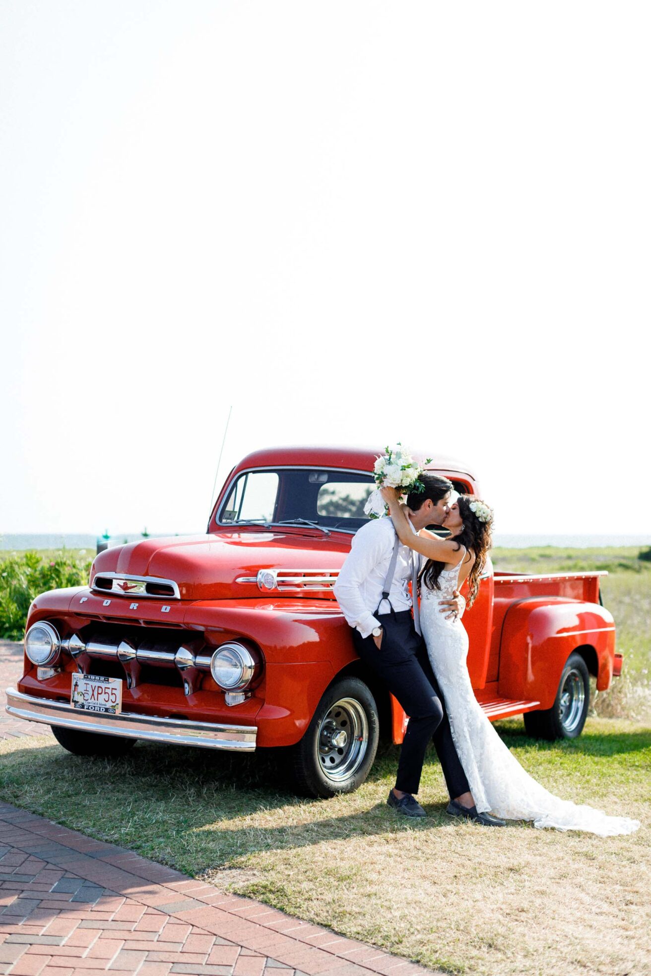 A newly wed couple sharing a kiss next to a red car.